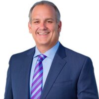 Portrait of a professional man in a suit with a purple striped tie, smiling against a white background.