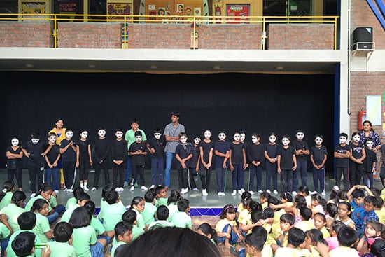 School children performing a mime act on stage wearing black outfits and painted white faces.