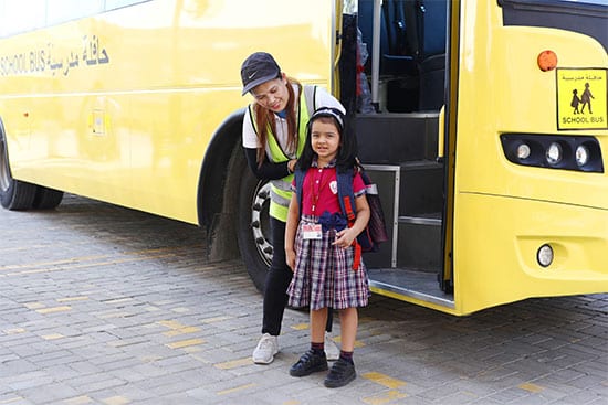 A schoolgirl posing with an adult near a school bus after arriving at school A child wearing a school uniform smiles while standing next to a school bus with a woman beside her, likely a caregiver or teacher