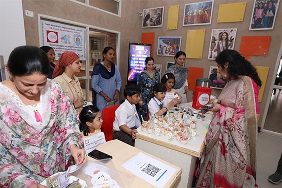 A group of adults and children are gathered at a table, participating in an event with sweets and donations being exchanged. The children, dressed in school uniforms, are helping to arrange or manage items, while adults, some holding money, observe and interact