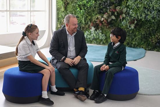 A meeting between a man and two students in a school environment A man sitting with two children, one girl and one boy, on blue seating cushions in a modern educational setting with greenery in the background
