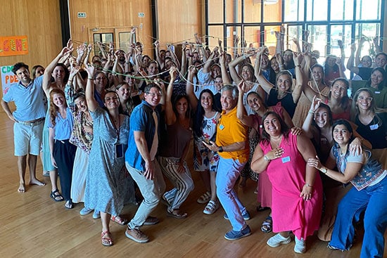 A vibrant group of people participating in a fun and collaborative event indoors Group of people standing together in a large indoor space, holding up colourful string decorations and smiling at the camera