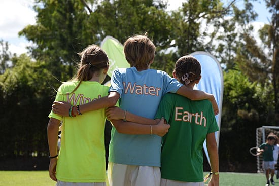 Children in themed shirts representing the elements Wind, Water, and Earth at an outdoor event Three children standing together with their arms around each other, wearing brightly coloured shirts labelled Wind, Water, and Earth