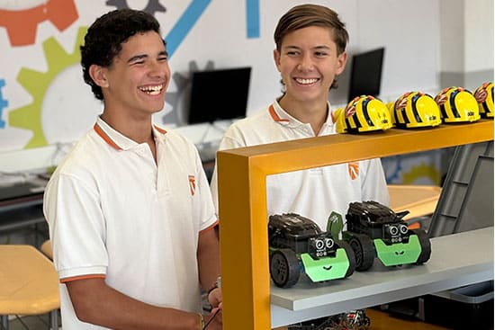 Students showcasing robots in a technology classroom Two smiling students standing next to a shelf displaying robotic cars and helmets in a classroom setting