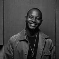 Theron 'Neff-U' Feemster Smiling man wearing a jacket and necklace, standing against a dark background.