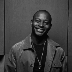 Smiling man wearing a jacket and necklace, standing against a dark background.