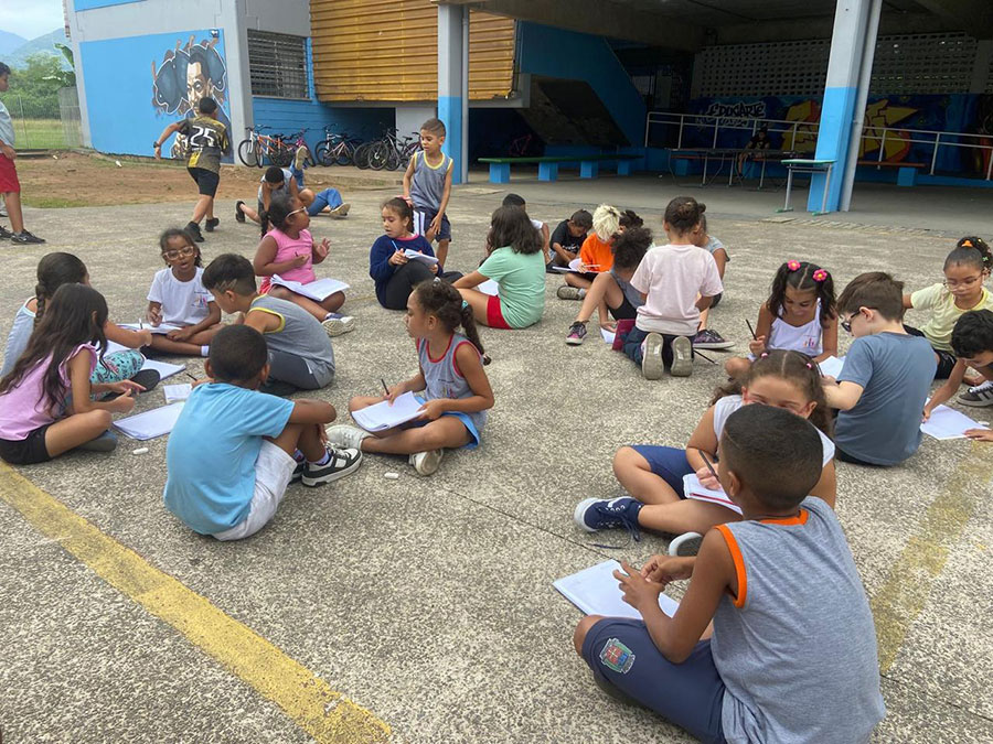 Children sitting in groups outside, writing and sharing ideas together.