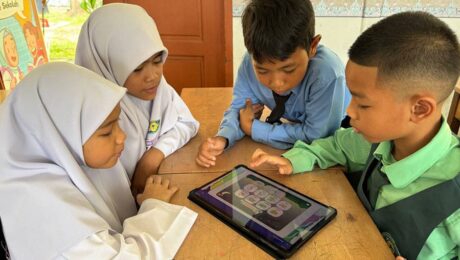 Children sitting together at a school desk using a tablet for learning activities.