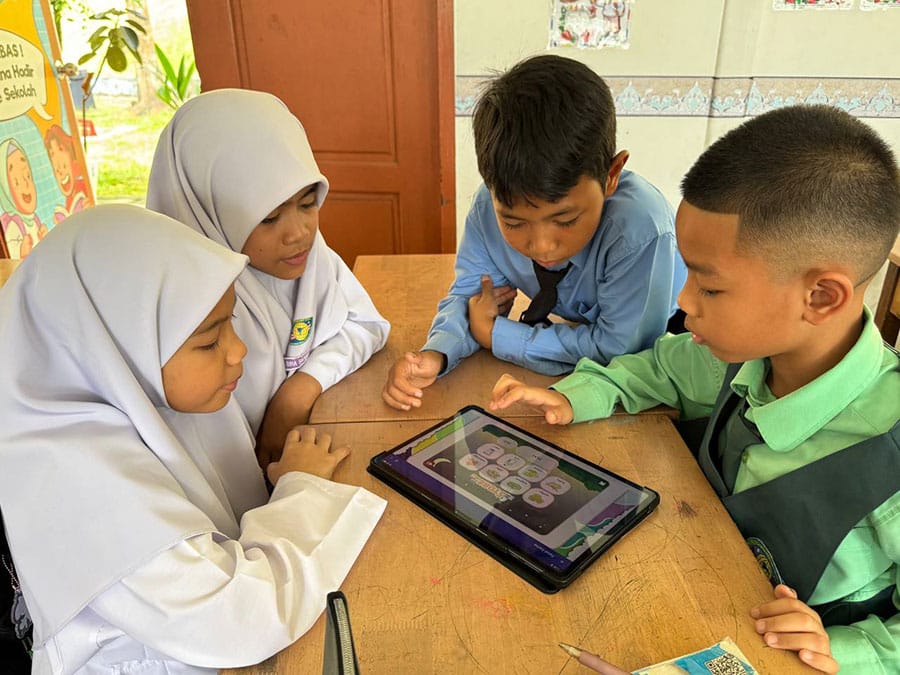 Children sitting together at a school desk using a tablet for learning activities.
