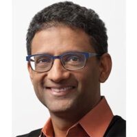 Ben Gomes Smiling person with short curly hair and glasses wearing an orange shirt and dark sweater