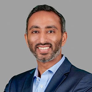 A man in a navy blue suit smiling and posing against a gray background.