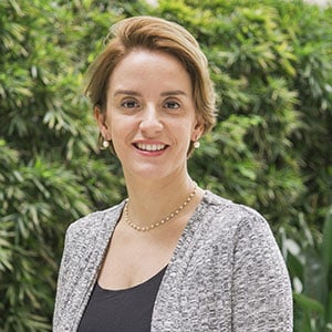A woman in a gray blazer smiling while standing outdoors with greenery in the background.