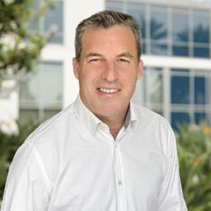 A man in a white shirt smiling outdoors with a modern building in the background.