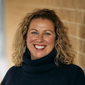A woman with curly hair wearing a dark turtleneck sweater, smiling while standing by a brick wall.