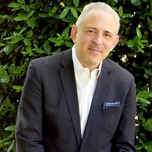 Portrait of David Barth, man in a dark suit standing outdoors with greenery behind