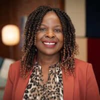 Eunice Ogolo Smiling woman with curly hair wearing a patterned blouse and blazer indoors