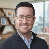 Smiling person with short dark hair and glasses wearing a zip-up sweater in an office setting
