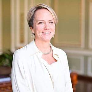 A woman with short hair wearing a light-colored blouse, smiling indoors in a well-lit room.