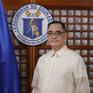A man wearing traditional Filipino attire standing beside a flag and the seal of the Department of Education of the Philippines.