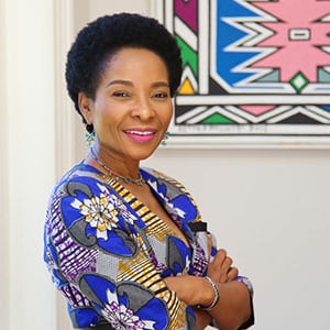 A woman in a colorful patterned outfit smiling with arms crossed, standing in front of vibrant artwork.