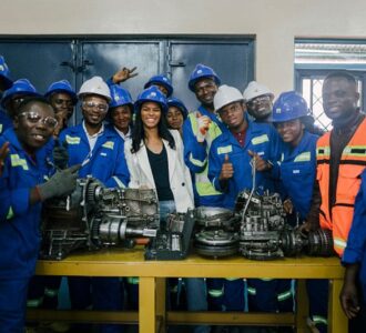 Group of trainees and instructors in blue overalls and safety helmets gathered around mechanical parts in a workshop.