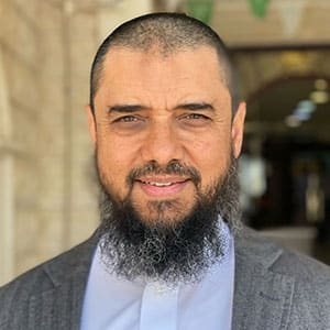 Portrait of a man with a trimmed beard wearing a suit jacket and white shirt, smiling outdoors.