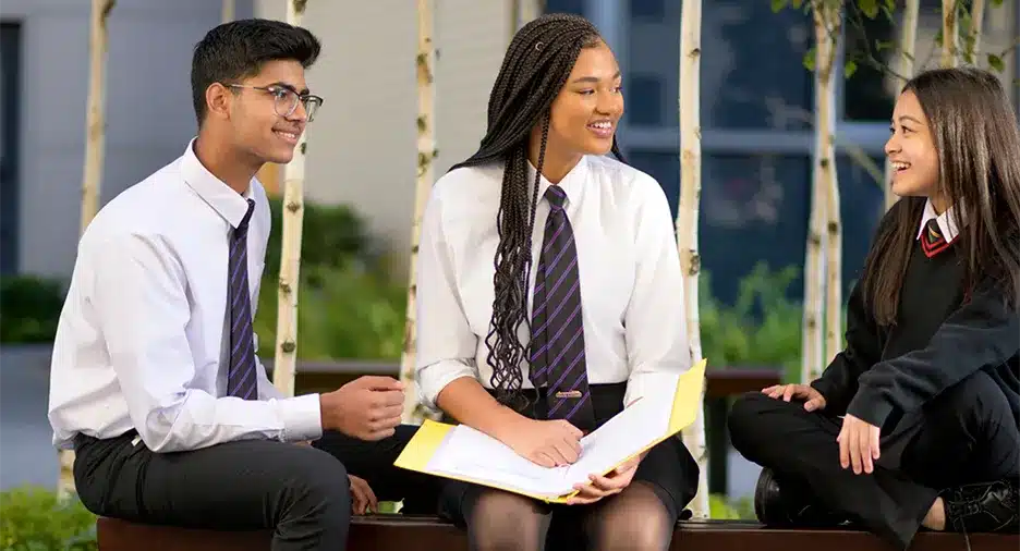 Three secondary school students in uniform sitting on a bench outdoors, discussing schoolwork and smiling together