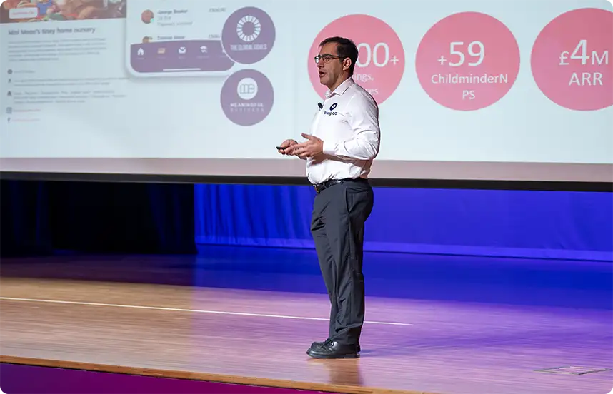 Business speaker delivering a presentation on stage with a large projected screen displaying company metrics and statistics
