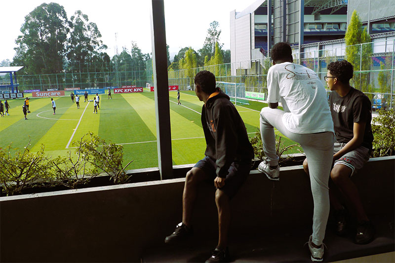 Students watching a football match from an indoor viewing area overlooking a sports field
